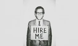 A man in a suit and glasses wearing a sign that says "hire me" 
