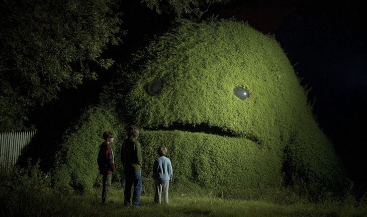 a large green hedge with eyes looking at young people 